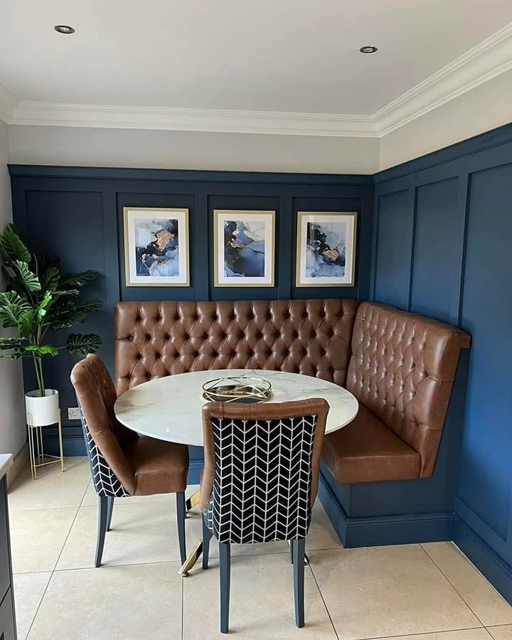 Dining nook with brown tufted banquette, round table, patterned chairs, framed art, plant and blue panelled walls