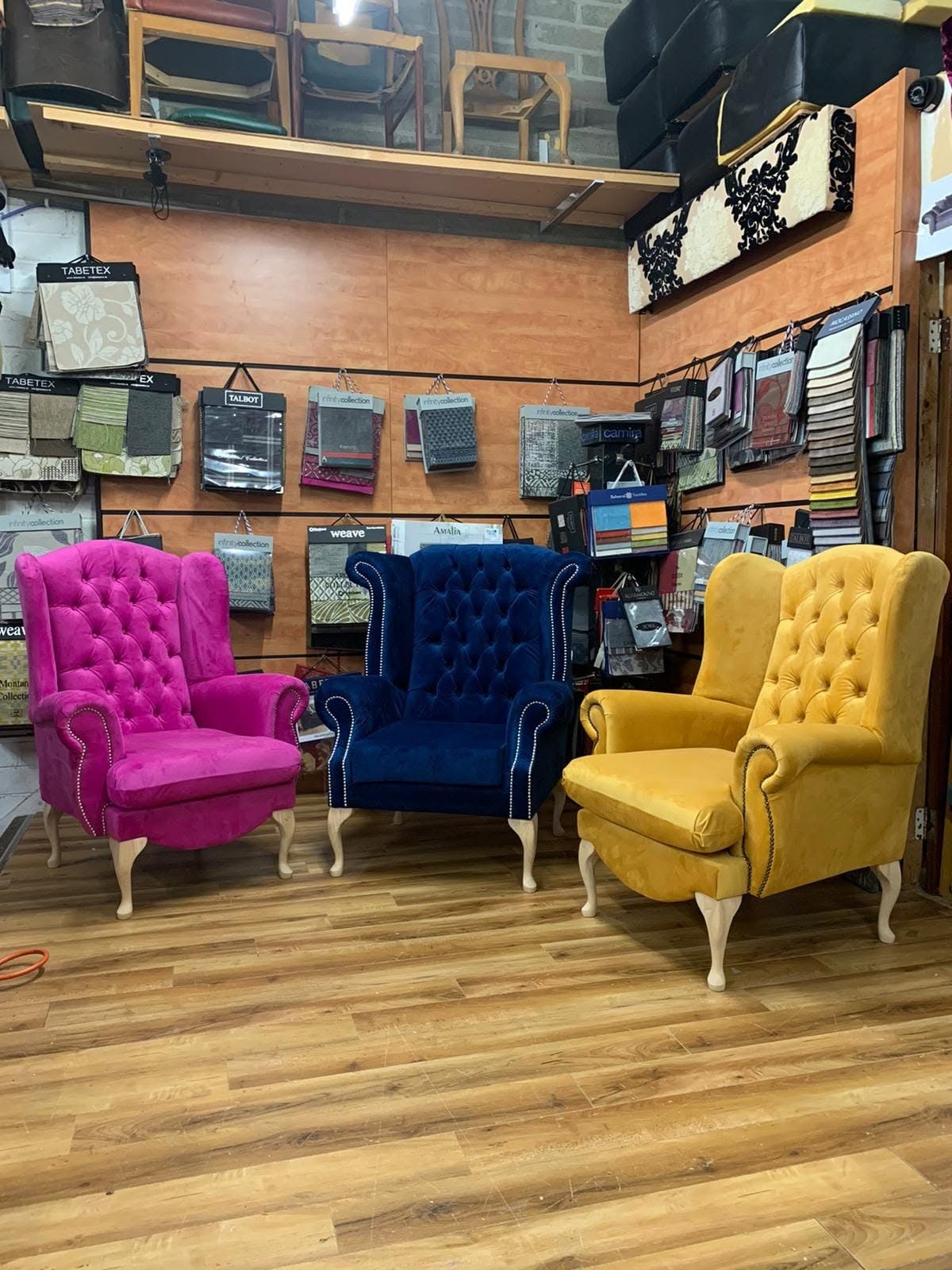 Three tufted wingback chairs in pink, blue and yellow in front of the fabric sample wall